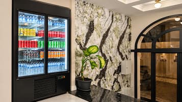 A modern lobby with a beverage refrigerator stocked with various drinks, a marble-patterned wall, and a potted plant.