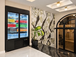 A modern lobby with a beverage refrigerator stocked with various drinks, a marble-patterned wall, and a potted plant. - Istanbul Efes Hotel (Istanbul)
