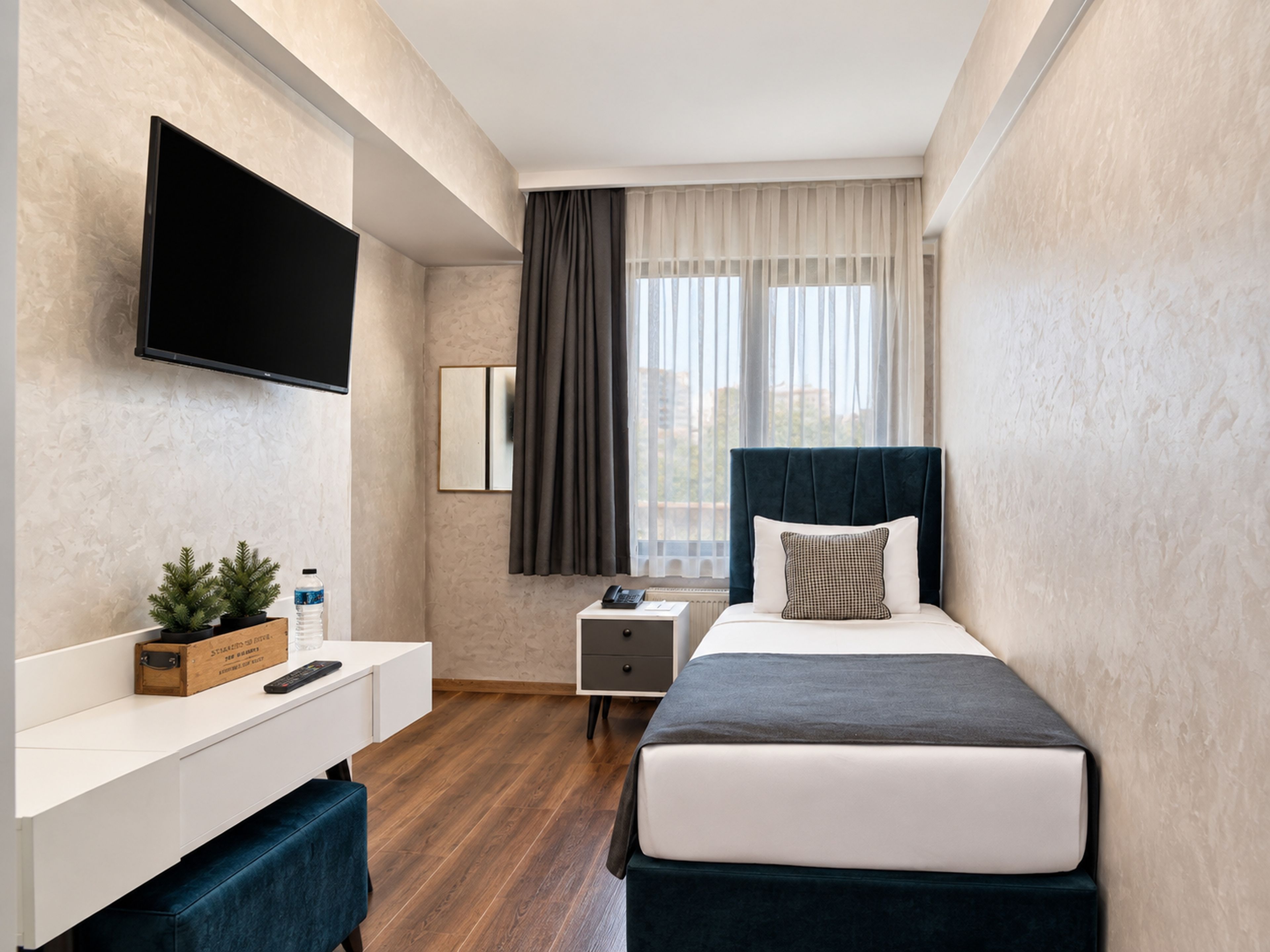 A hotel room with a bed, a TV mounted on the wall, a bedside table, a nightstand, a small plant, and a window with curtains.