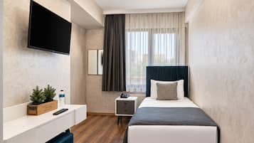 A hotel room with a bed, a TV mounted on the wall, a bedside table, a nightstand, a small plant, and a window with curtains.