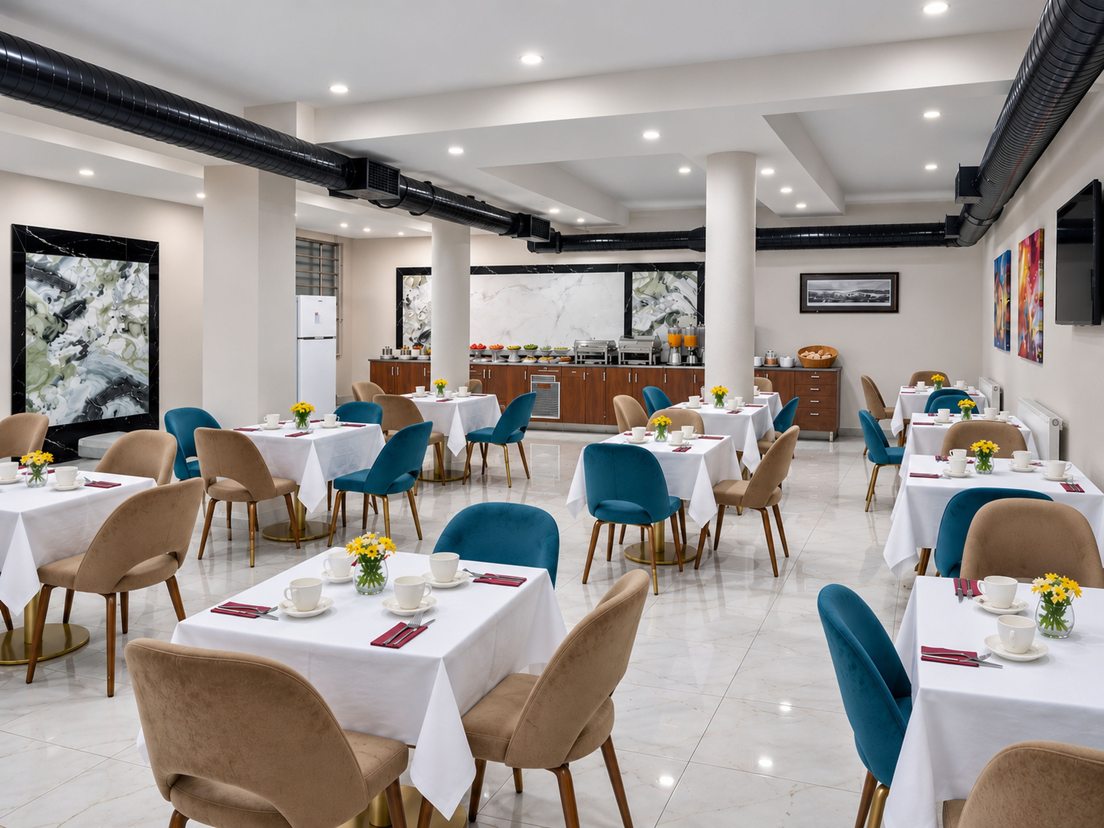 A hotel dining area with multiple tables and chairs, a kitchen in the background, and artwork on the walls.