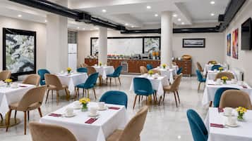A hotel dining area with multiple tables and chairs, a kitchen in the background, and artwork on the walls.