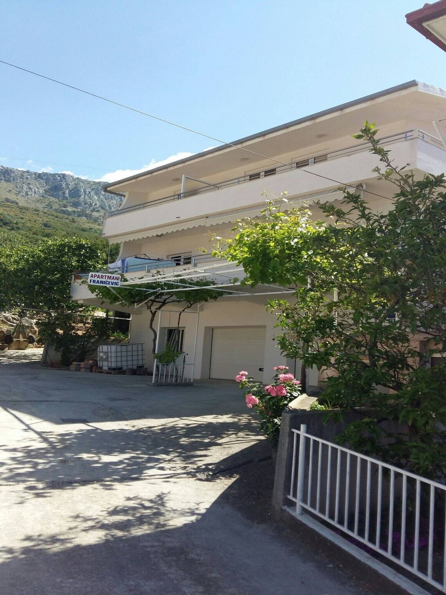 A two-story building with a sign indicating it is an apartment. There is a garage door, a small garden with pink flowers, and a mountain in the background.
