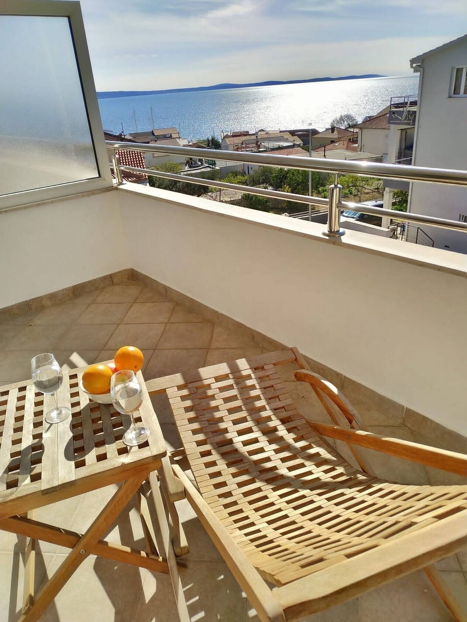 A balcony with a view of the sea, a table with oranges and glasses, and a wooden chair.