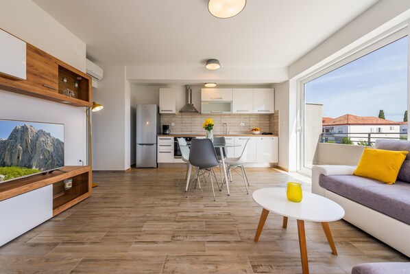 A modern living room with a wooden floor, a dining area with a glass table, and a kitchen in the background. - Modern two bedroom apartment with shared pool (Zadar)