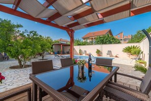 An outdoor patio with a glass table, wicker chairs, and a pool in the background. - Modern two bedroom apartment with shared pool (Zadar)