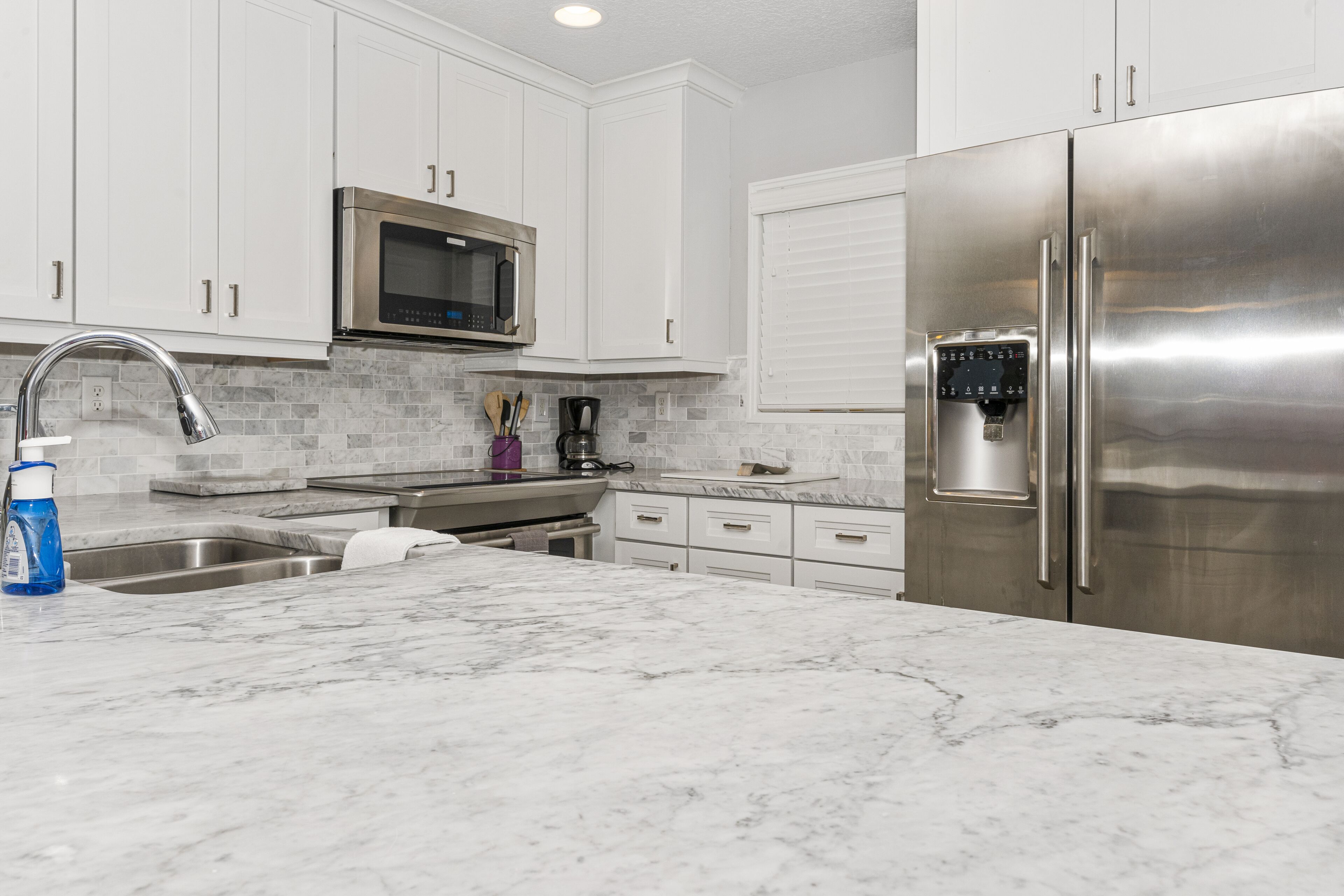 A modern kitchen with white cabinets, a stainless steel refrigerator, a microwave, and a marble countertop.