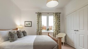 Um quarto com cama, mesa de cabeceira com abajur, cadeira, janela com cortinas e porta.