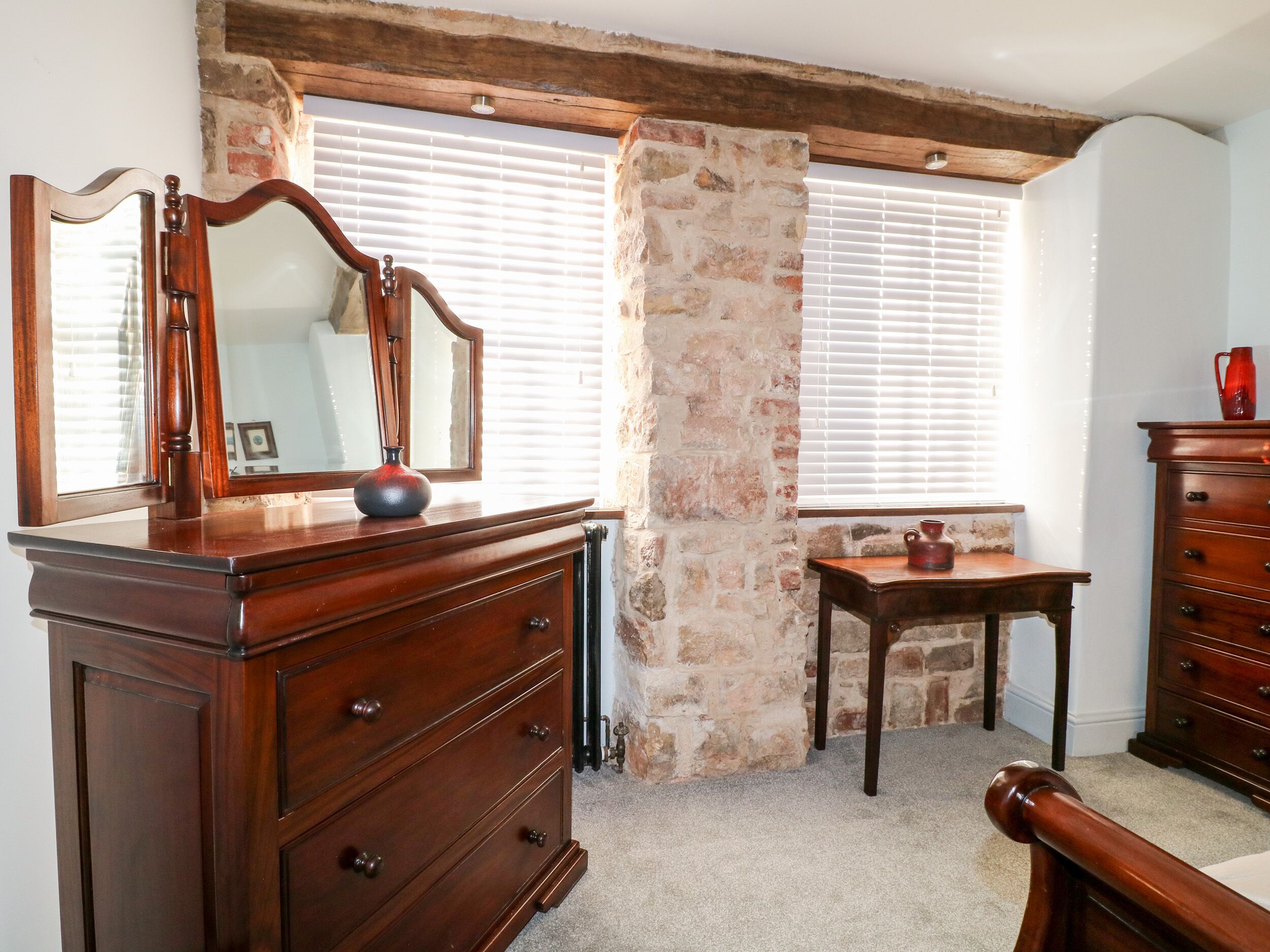 Une pièce avec une commode en bois, un miroir, une petite table sur laquelle est posé un vase, et un mur en pierre.