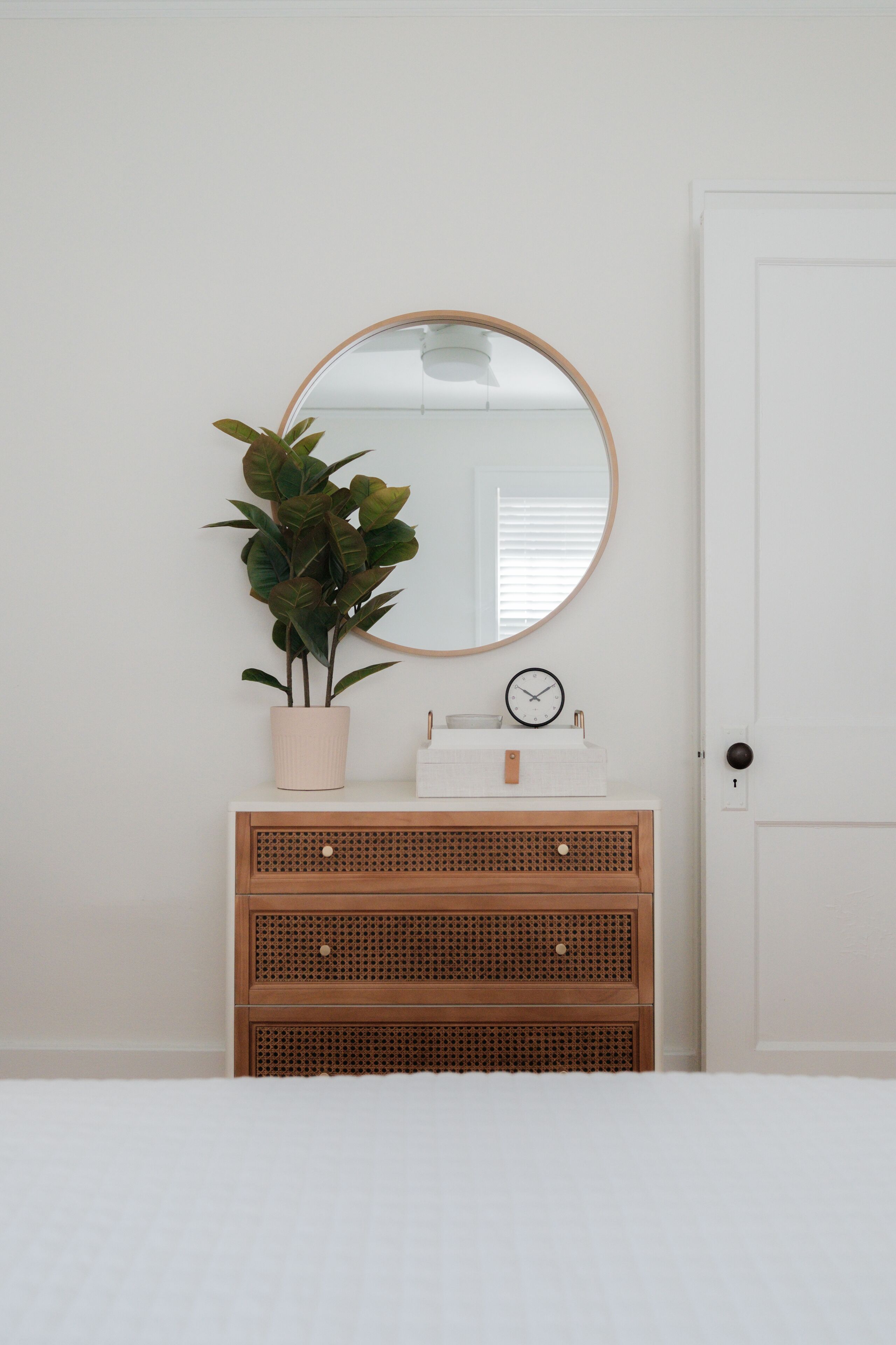Una camera da letto con un letto bianco, una cassettiera di legno con cassetti, uno specchio rotondo, una pianta in vaso e una sveglia sulla cassettiera.
