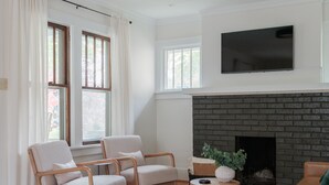 A modern living room with a fireplace, wooden flooring, and a flat-screen TV mounted on the wall. - Stylish Family/Pet Friendly Cottage in Coveted Clark Park with Fenced In Yard  (Champaign)
