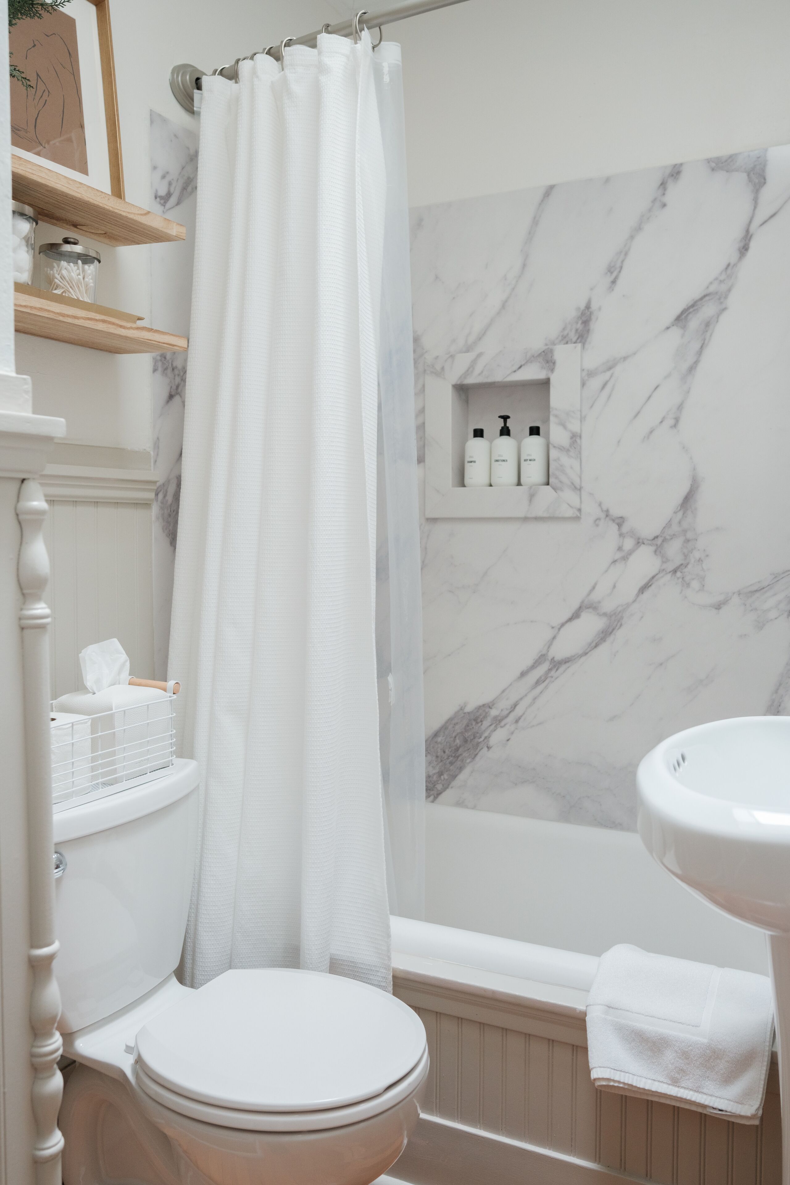 A bathroom with a toilet, a bathtub, a shower curtain, marble walls, and a shelf with toiletries.