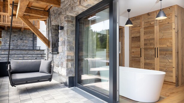 A modern interior with a stone wall, a swing sofa, a glass door leading outside, and wooden cabinets. - Superior Chalet for 8 people with sauna (Turrach)