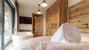 A modern bedroom with a large bed, a bathtub, wooden wardrobe, and a TV mounted on the wall. - Superior Chalet for 8 people with sauna (Turrach)