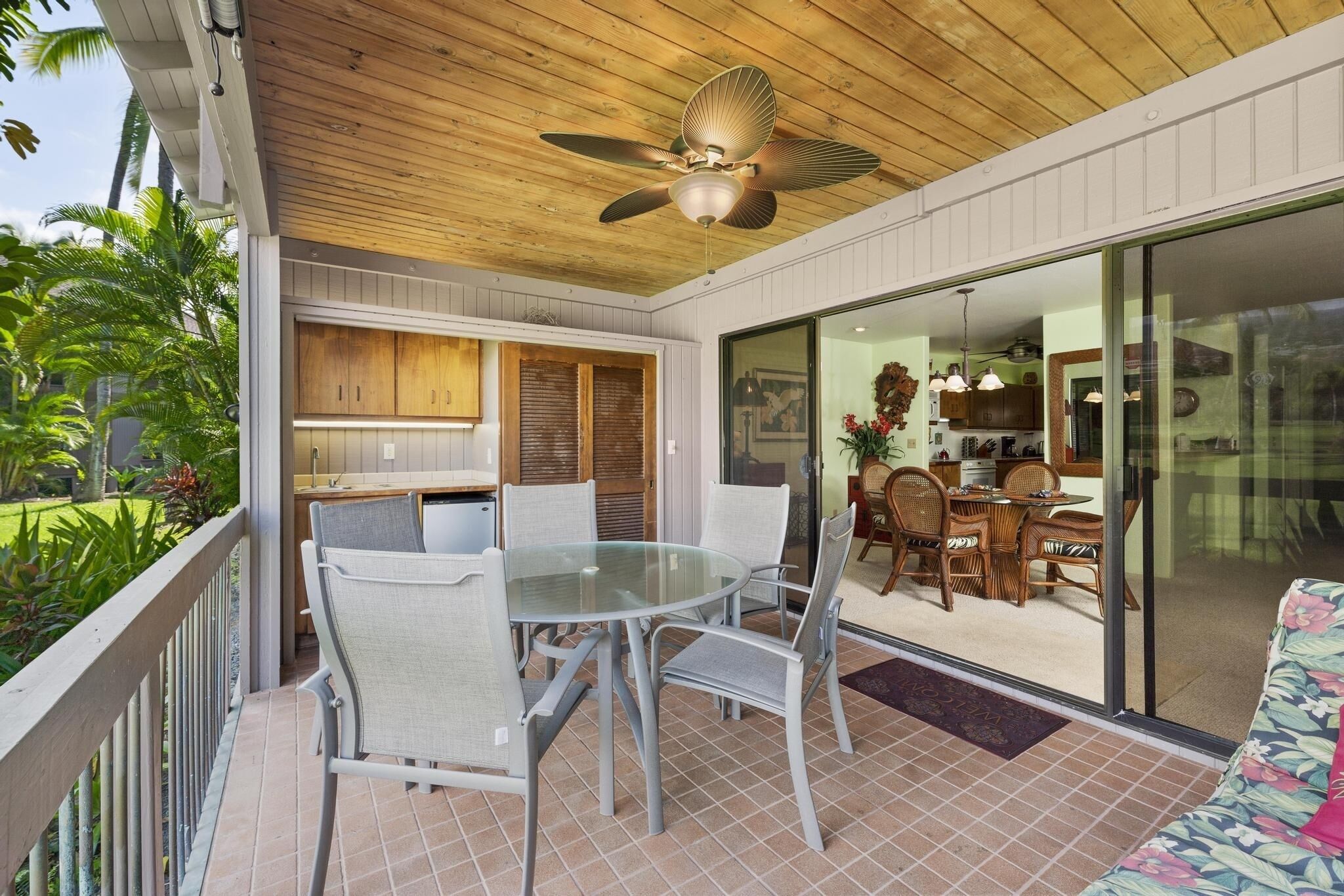 A covered outdoor dining area with a glass table and chairs, a ceiling fan, and a view of the adjacent kitchen and living space.