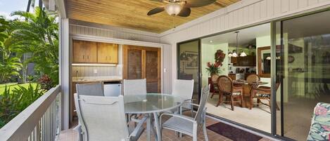 A covered outdoor dining area with a glass table and chairs, a ceiling fan, and a view of the adjacent kitchen and living space.