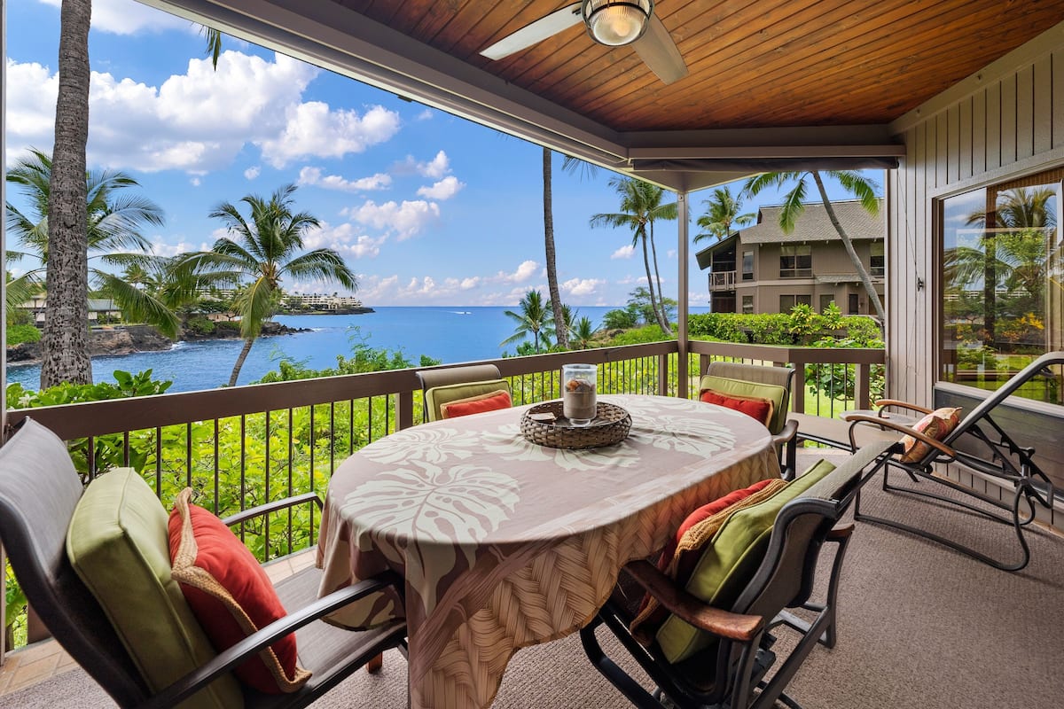 A covered outdoor seating area with a table and chairs, overlooking a scenic ocean view with palm trees and a building in the background.