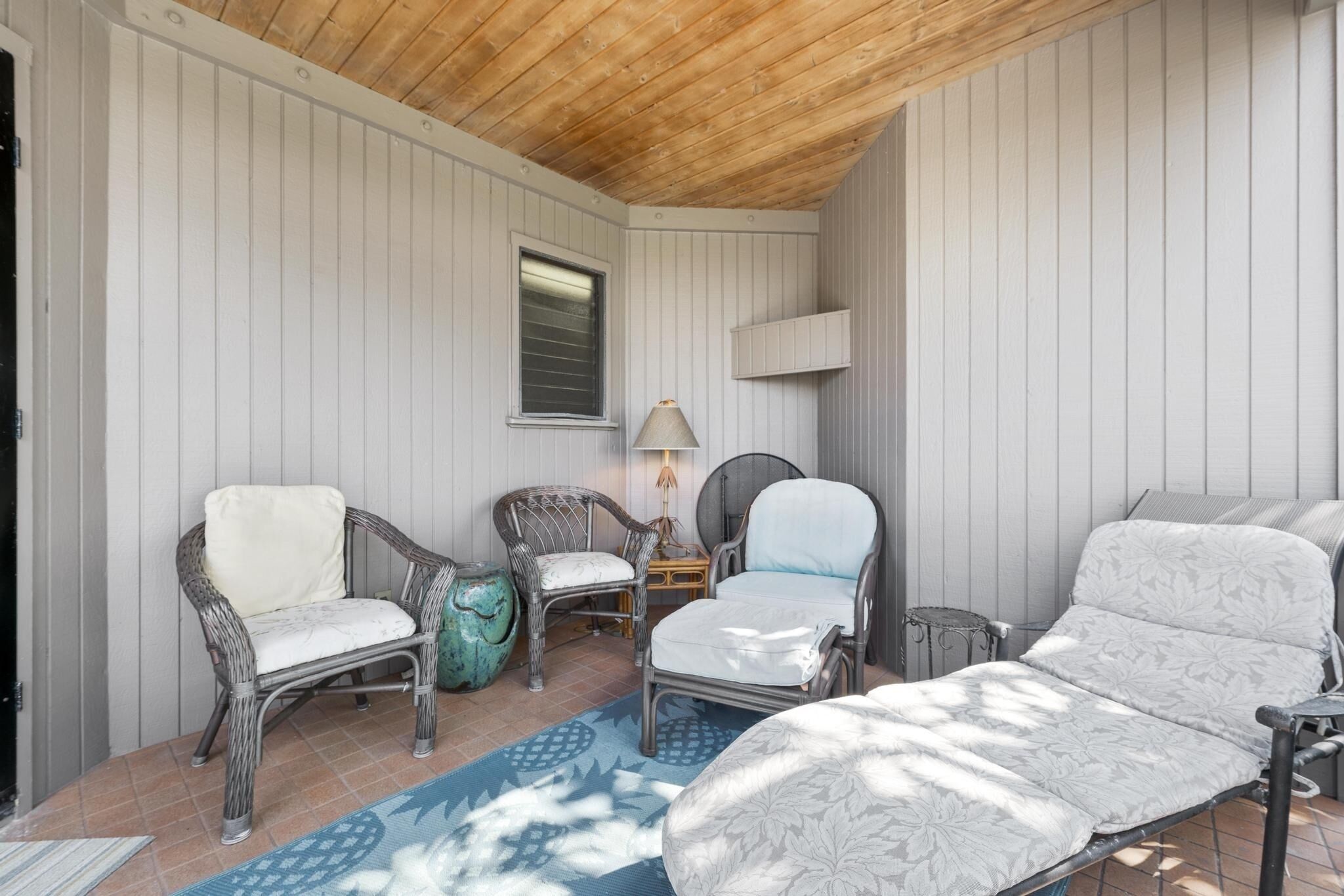 A cozy room with a wooden ceiling, white paneled walls, and a large window. There are two wicker chairs with cushions, a small table with a lamp, and a sofa with a patterned cover.