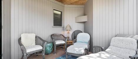 A cozy room with a wooden ceiling, white paneled walls, and a large window. There are two wicker chairs with cushions, a small table with a lamp, and a sofa with a patterned cover.