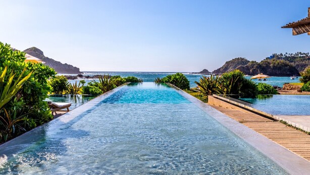 A long, narrow infinity pool with clear blue water, surrounded by lush greenery and overlooking a coastal view. - Condo 223 El Careyes Club & Residences (Costa Careyes)