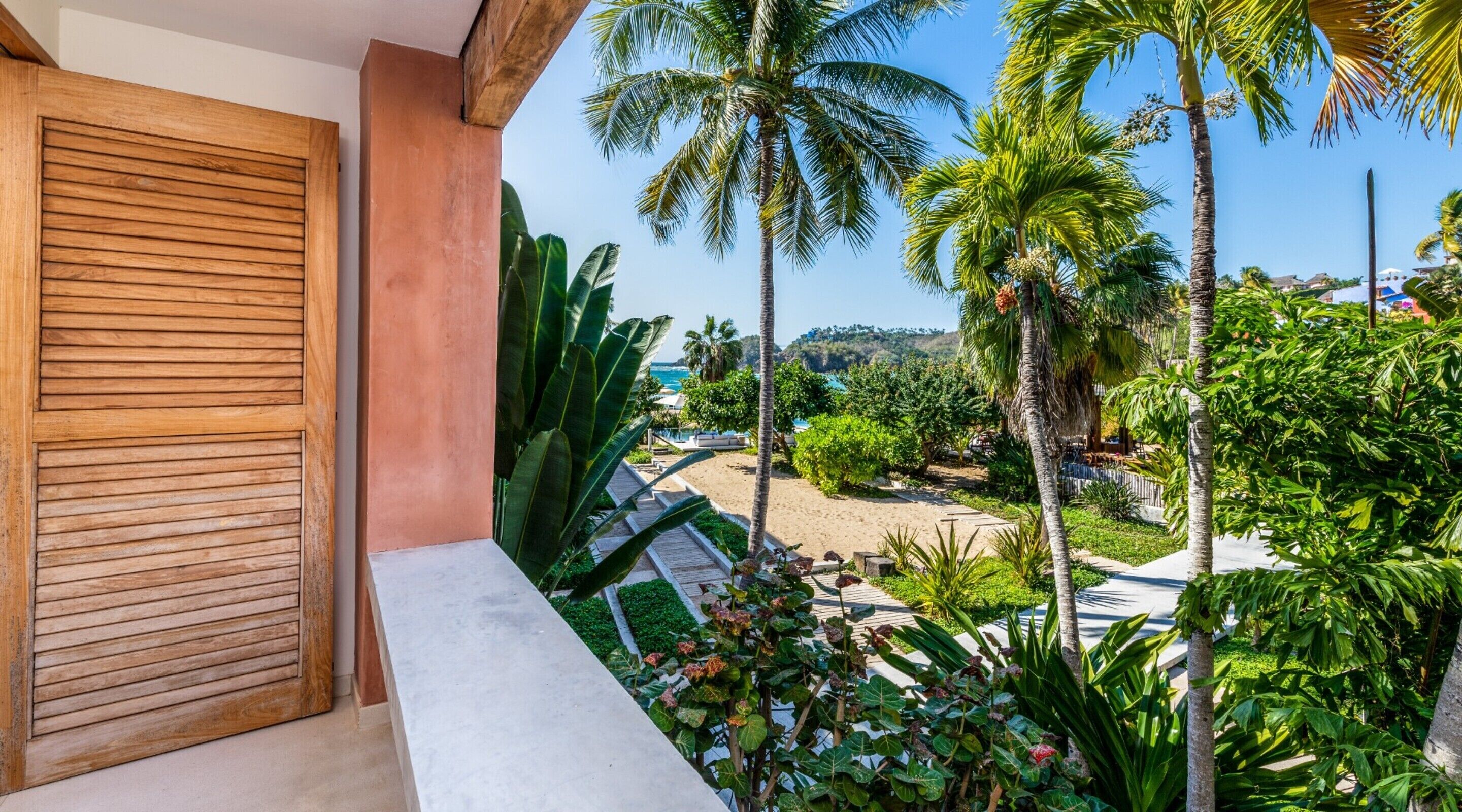 A balcony with a wooden shutter, a stone ledge, and a view of a beach with palm trees and a clear sky.