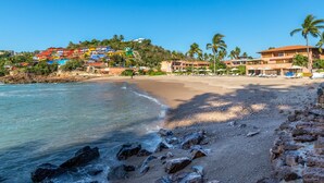 A beach with a rocky shoreline, palm trees, and colorful buildings in the background. - Condo 223 El Careyes Club & Residences (Costa Careyes)
