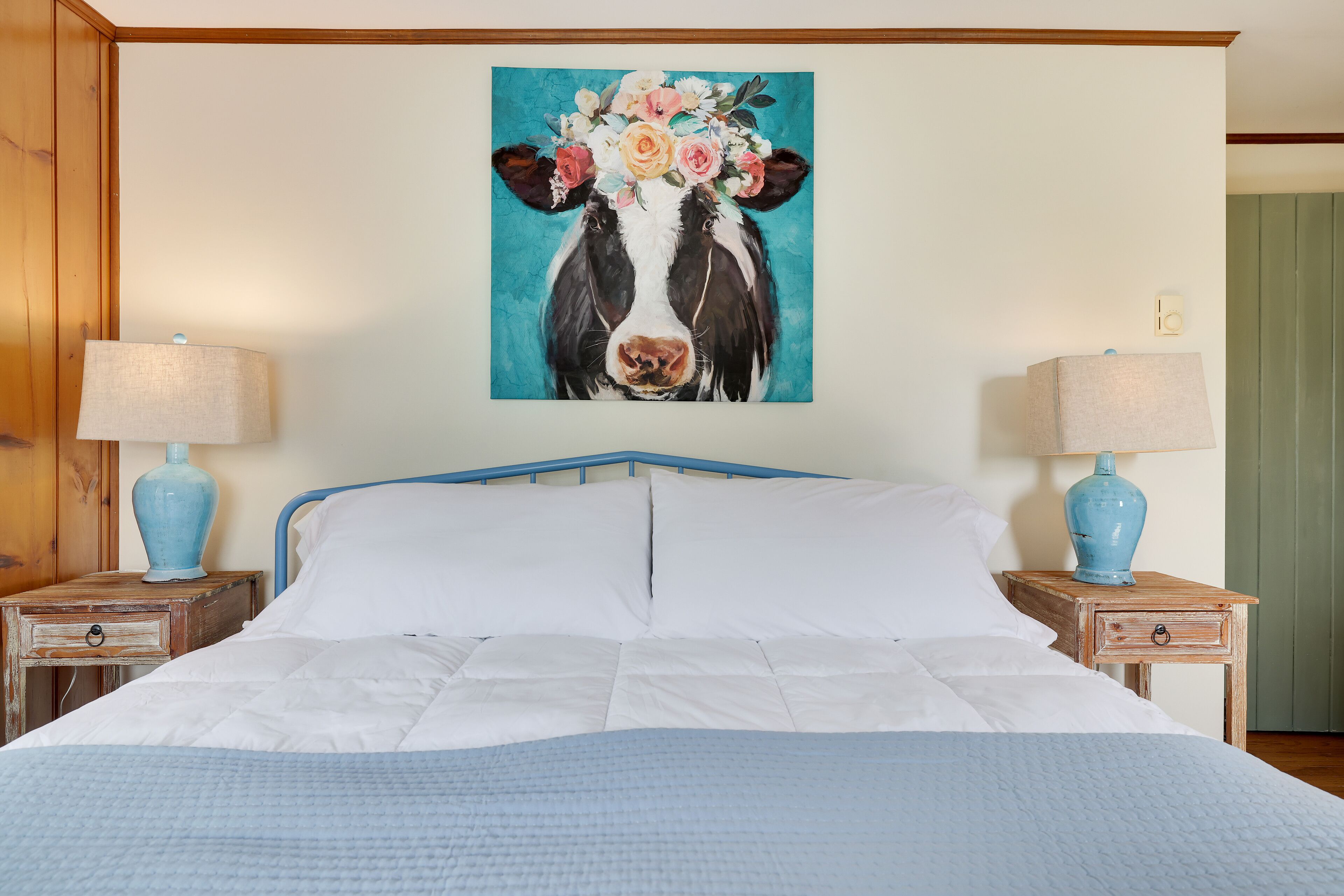 Une chambre à coucher avec un lit, deux tables de chevet avec des lampes et un tableau mural représentant une vache entourée de fleurs.