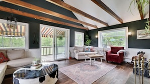Un salón espacioso con techo alto, vigas de madera, ventanas amplias y una zona de estar acogedora con un sillón rojo y un sofá blanco.