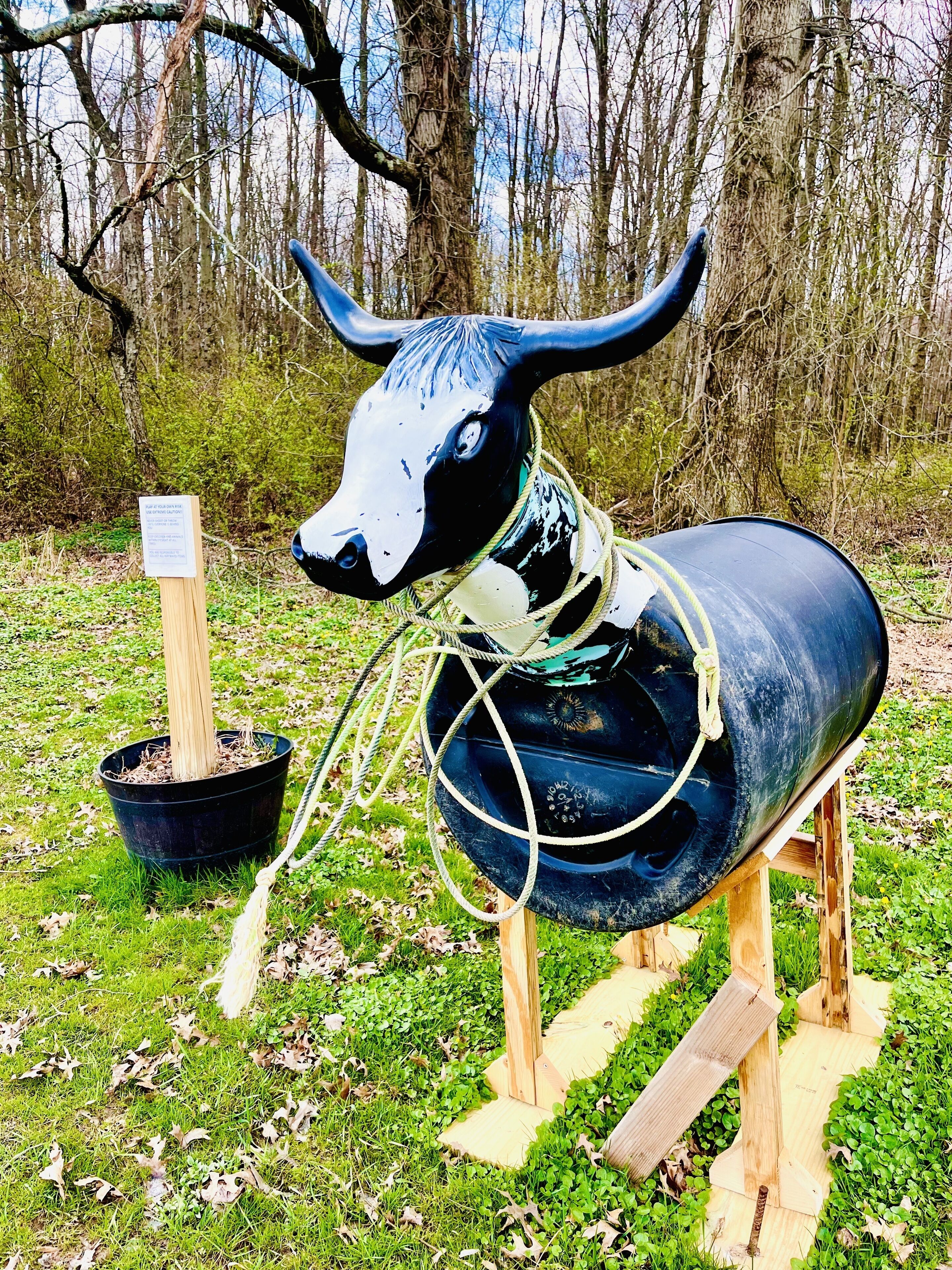 A sculpture of a bull with a rope around its neck, standing on wooden legs, in a grassy area with trees in the background.