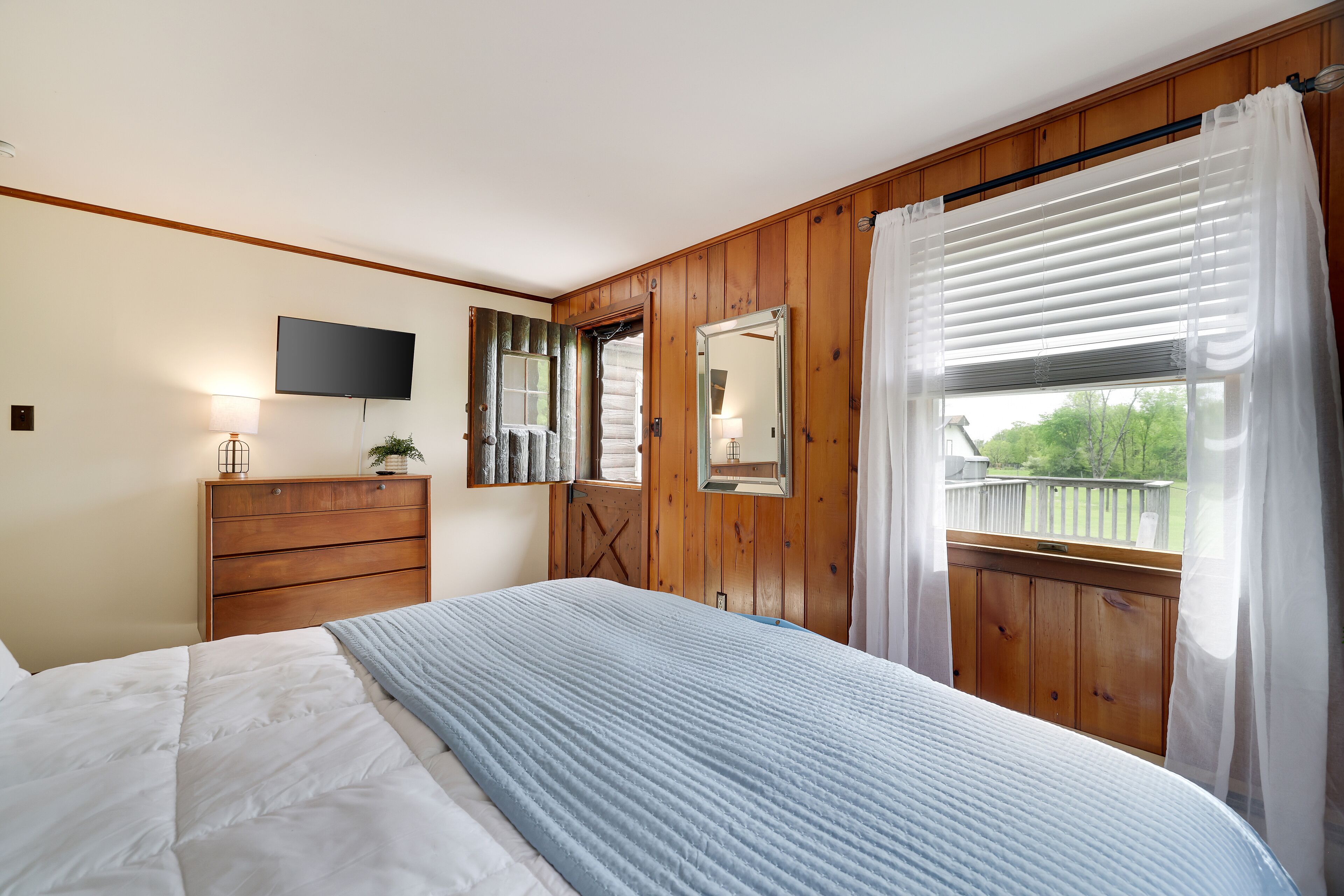 A bedroom with a bed, a dresser, a TV, a mirror, and a window with curtains.