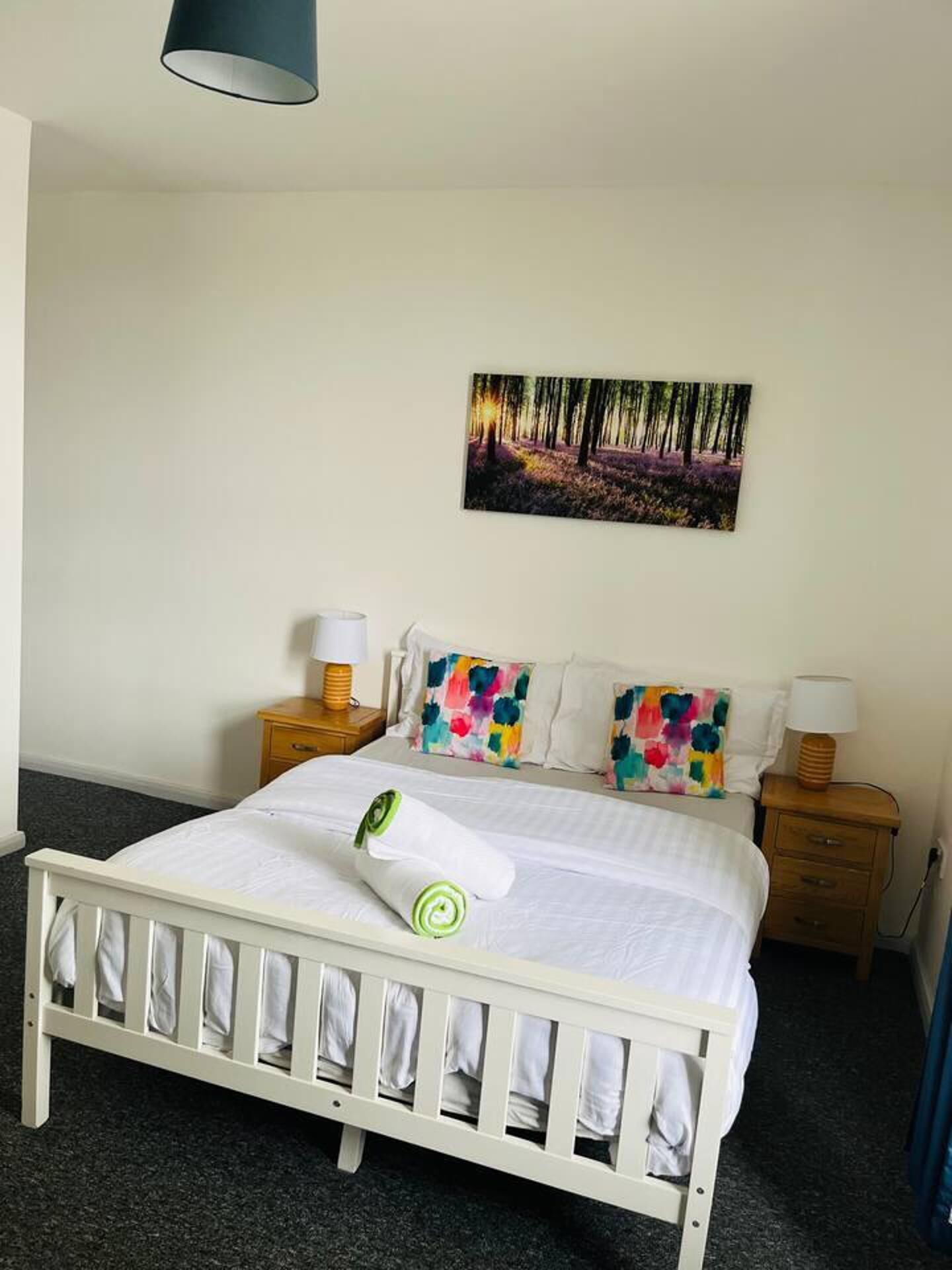 Ein Schlafzimmer mit einem weißen Bett, bunten Kissen, Wandbild mit Waldmotiv und zwei Nachttischlampen.