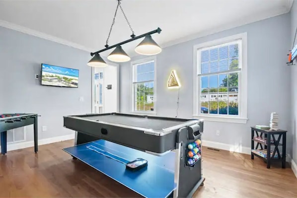 A pool table with a blue felt surface, a foosball table, and a billiards table with a black felt surface. There are two pendant lights above the pool table.