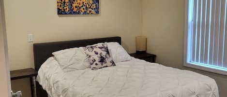 Une chambre à coucher avec un lit, une table de chevet, un tableau, une fenêtre avec des stores et une porte.