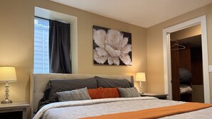 Un dormitorio con una cama, dos mesitas de noche, un ventanal con cortinas, un cuadro en la pared y un vestidor.