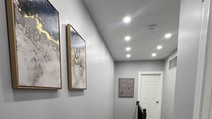 Ein Flur mit grauen Wänden, Holzdielen und eingelassener Beleuchtung. An der linken Wand befinden sich zwei gerahmte Kunstwerke und ein Tür am Ende.