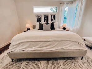 Een slaapkamer met een groot bed, twee nachtkastjes, een raam met gordijnen en kunstwerken in lijsten aan de muur.
