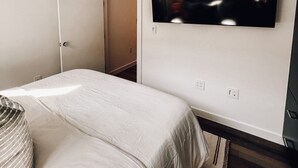 Un dormitorio con cama, ventilador de techo, televisor montado en la pared y puerta que da a otra habitación.
