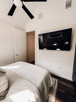 A bedroom with a bed, a ceiling fan, a television mounted on the wall, and a door leading to another room.