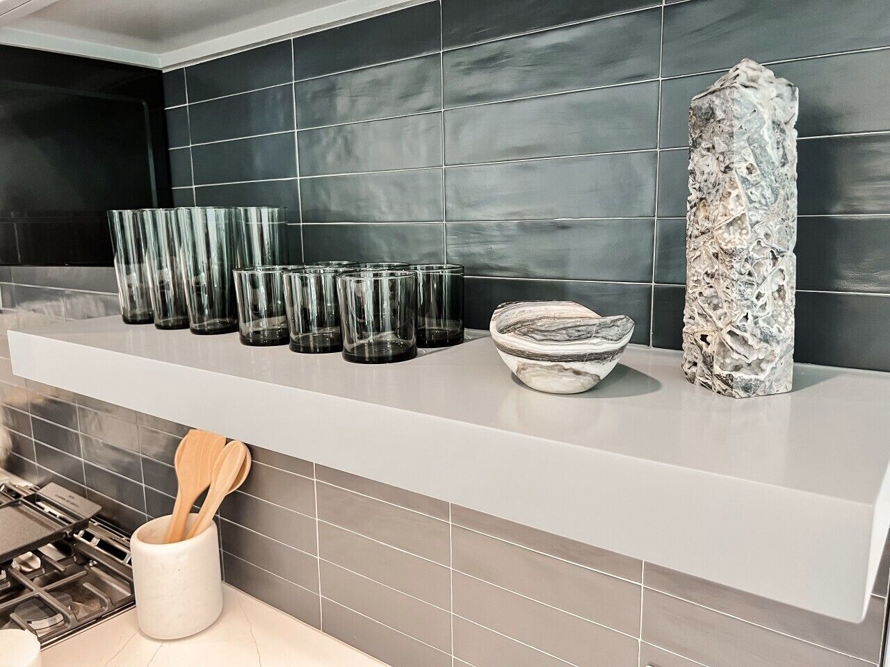A kitchen counter with a white shelf holding several glass tumblers, a marble bowl, and a textured vase.
