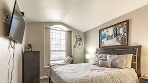 Een slaapkamer met een groot bed, een aan de muur bevestigde televisie, een raam met zonwering en een schilderij boven het bed.