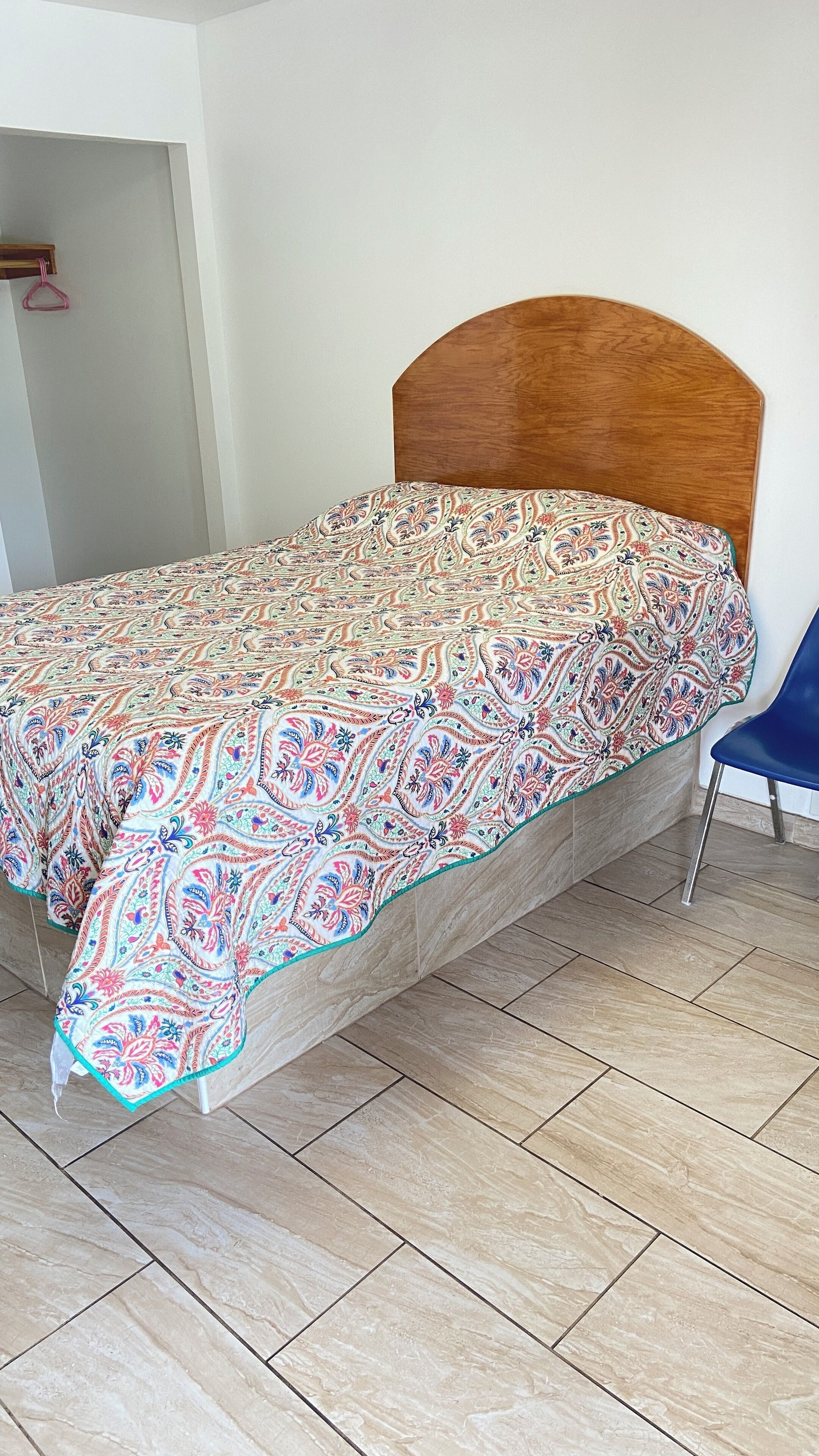 Een bed met houten hoofdeinde, een bed met een decoratief laken en een blauwe stoel in een kamer met betegelde vloer.