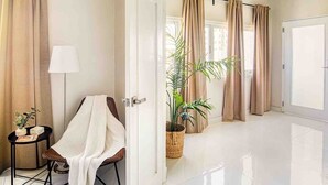 Habitación de hotel con piso de baldosas blancas, una silla con una toalla blanca, una mesita con una lámpara, una planta en maceta y cortinas color beige.