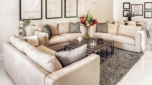 Una sala de estar moderna con un sofá seccional color beige, una mesa de centro y una alfombra. Hay obras de arte abstractas enmarcadas en la pared y un jarrón con flores sobre la mesa de centro.