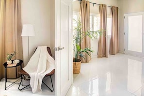 Habitación de hotel con piso de baldosas blancas, una silla con toalla blanca, una mesita con lámpara, una planta en maceta y cortinas color beige.
