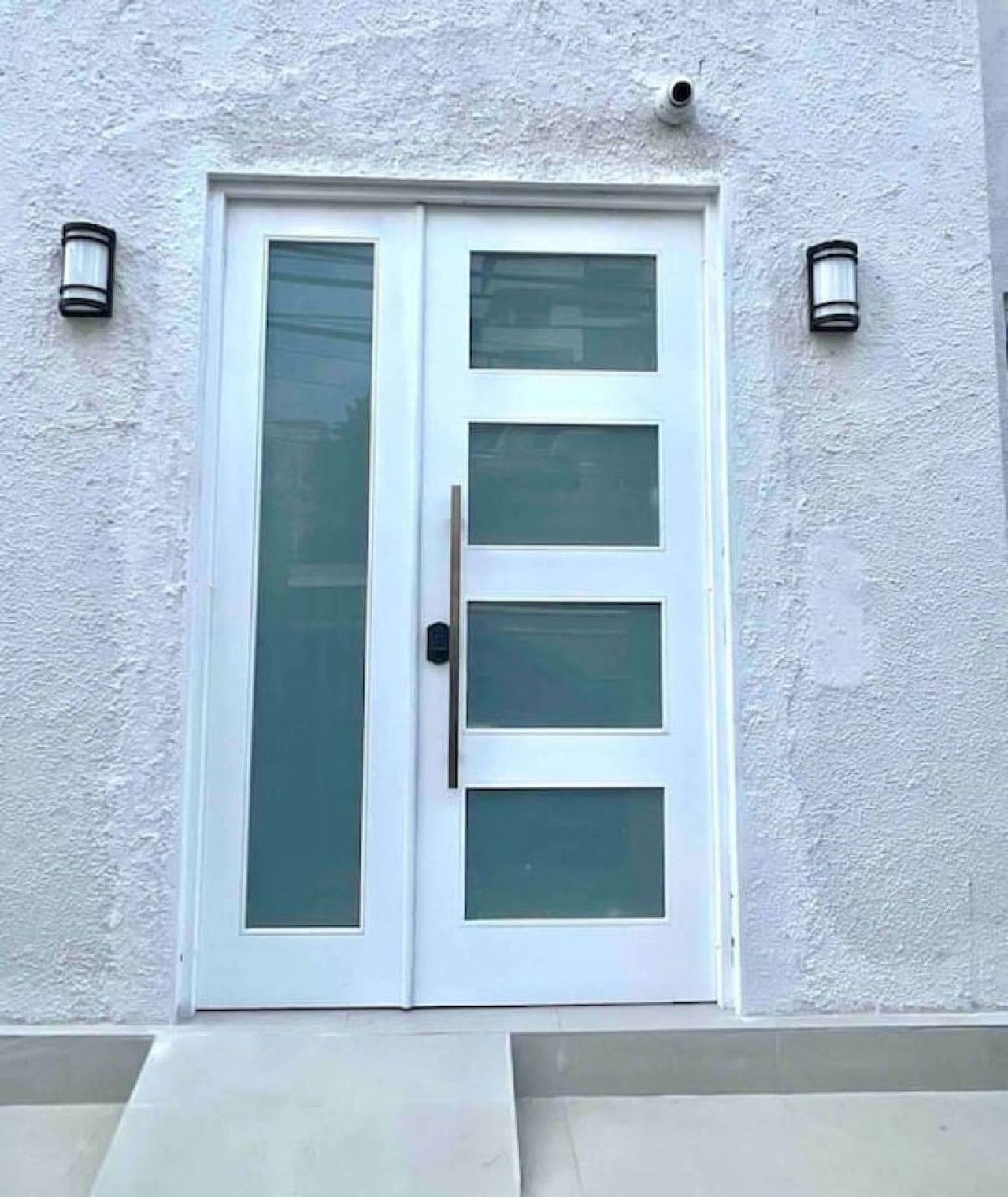 Una puerta blanca con un panel de vidrio vertical y una manija, flanqueada por dos lámparas fijadas a la pared.