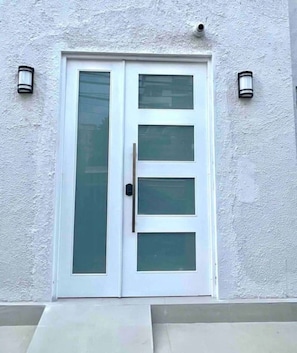 Una puerta blanca con un panel de vidrio vertical y una manija, flanqueada por dos lámparas fijadas a la pared.