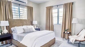 Un dormitorio moderno con una cama grande, dos mesitas de noche con lámparas, una silla y vistas al exterior a través de amplios ventanales.