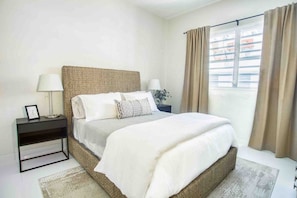 Un dormitorio con cama, mesitas de noche, una ventana con cortinas y vistas a edificios.