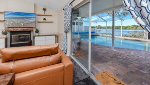 Una sala de estar con un sofá de cuero, un televisor de pantalla plana y una chimenea. Hay una puerta de vidrio que da a un área de piscina con vistas a un lago.