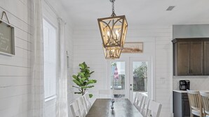 A dining room with a long table and white chairs, a hanging light fixture, and a view of the backyard through a door. -  Private Beach Access, Pool and Hot Tub! (Destin)